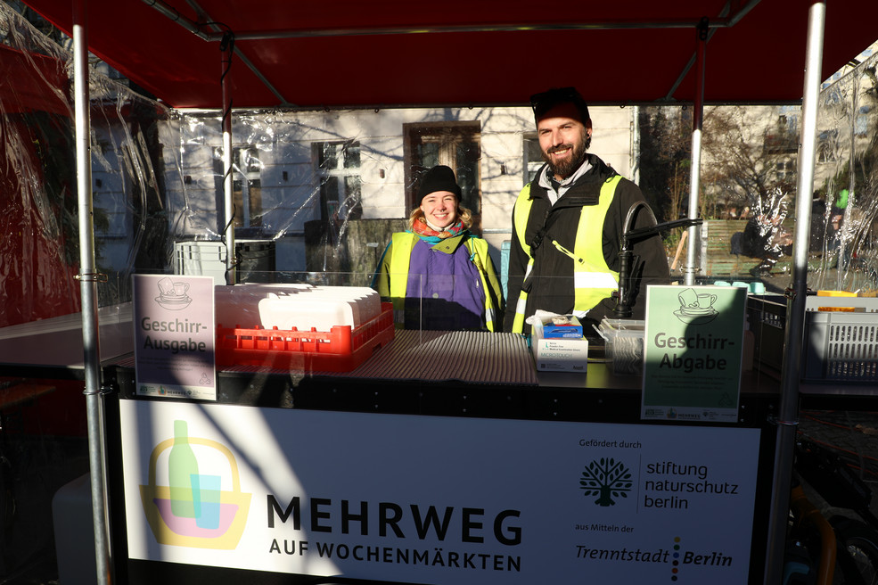 Zwei Personen stehen hinterm Spülmobil der GRÜNEN LIGA Förderprojekt Trenntstadt Mehrweg auf Wochenmärkten