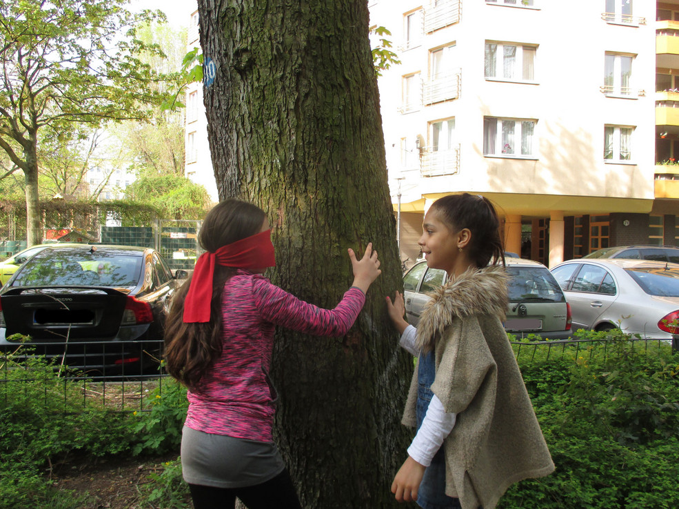 Zwei Mädchen tasten einen Baum mit ihren Händen ab. Das Mädchen auf der linken Seite hat ihre Augen verbunden.