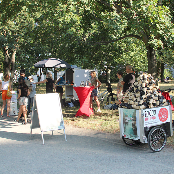 Viele Objekte und Menschen sind am Info-Stand eines Clean-up-Events zu sehen. Im Hintergrund ist der Park und Bäume.